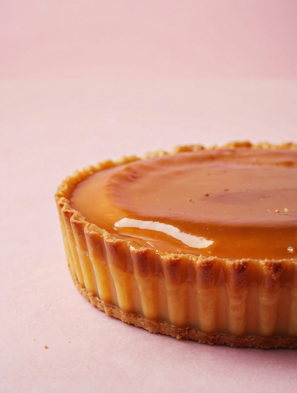 A close-up of a glossy caramel-topped tart with a golden pastry crust resting on a soft pink surface