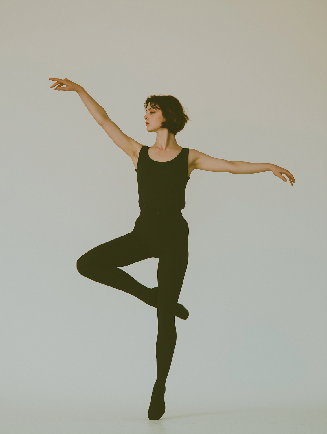 A dancer dressed in black balanced on one leg in mid-pose against a plain background