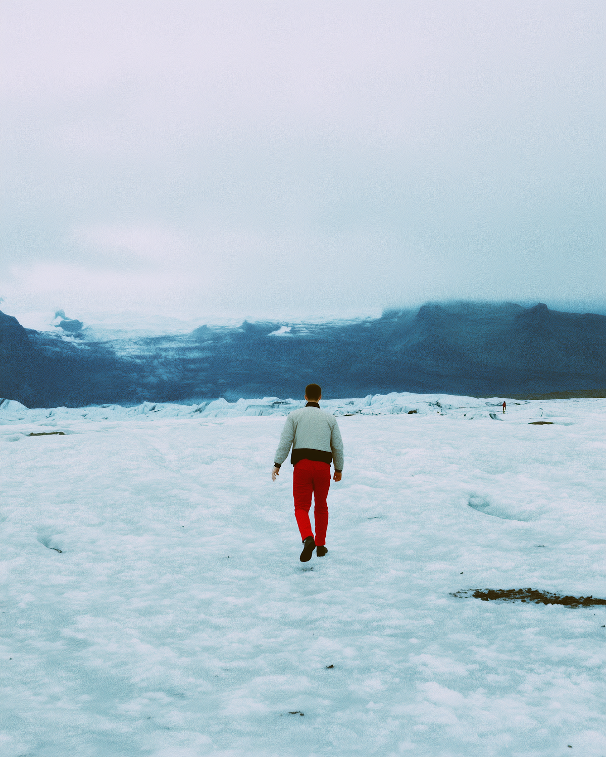 A lone walker in red trousers crossing a wide, empty snowfield with mountains rising behind