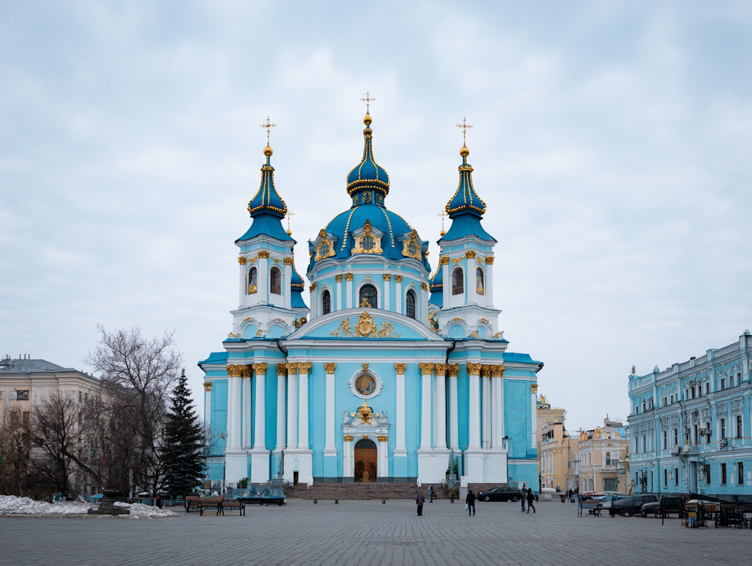 A large blue-domed church with gilded spires and white stonework rising above an open plaza under heavy cloud
